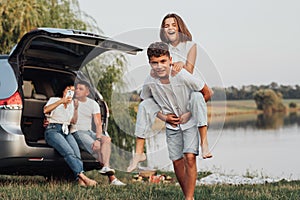 Two Happy Teenage Children Playing by the Lake While Their Parents Sitting in Trunk of the Minivan Car, Four Caucasian