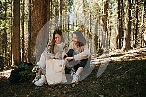 Two happy women in casual clothes sit in the woods and rest, look in the eco bag and rest