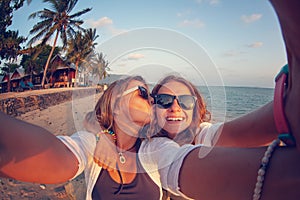 Two happy women girlfriends lesbian couple laughing while spending time on a tropical beach, summer vacation. Selfie and kiss