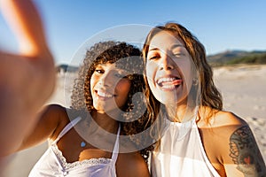 Two happy women multiracial best friends taking selfie on the beach with funny tongue faces wearing boho clothes. Tattooed girl