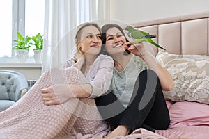 Two happy women sitting together on the bed with pet green parrot