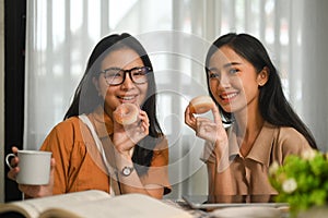 Two happy young asian female best friends eating donuts and enjoying free time together at home