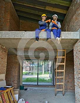 Two happy young builders in blue overalls and hard hats resting, sitting on the concrete floor, drinking coffee while