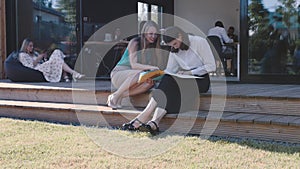 Two happy young female business partners sit on trendy office terrace outside, have coffee working together on sunny day