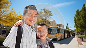 Two Happy Young Hispanic Brothers Wearing Backpacks Near a School Bus on Campus