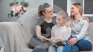 Two happy young smiling same sex mother reading book with their daughter at domestic interior