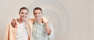Two happy young twin brothers hugging and smiling at camera while posing together isolated over beige background