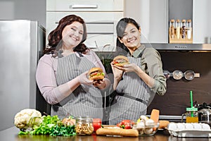 Two happy young women in aprons holding delicious burgers in their hands and looking at the camera in the kitchen