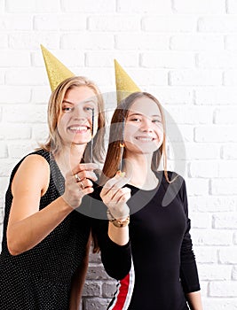 Two happy young women in birthday hats holding sparklers celebrating birthday, wearing golden birthday hats over white