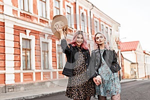 Two happy young women with lovely smiles with pink hair in stylish dresses in fashion jackets walk on road on street in city.