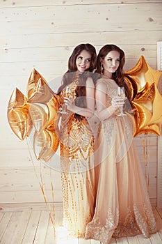 Two happy young women on party with glasses of champagne. Cheerful girls in long fashion dress by golden stars balloons over