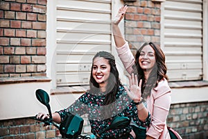 Two happy young women riding on motorbike together through city street and waving their friends