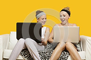 Two happy young women using laptop sitting on sofa against yellow background