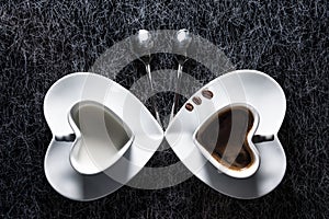 Two heart shaped cups with black coffee and milk pointing to each other, with three coffee beans