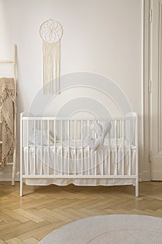 Two heart shaped pillows placed on baby crib standing in white room interior with macrame on wall and herringbone parquet