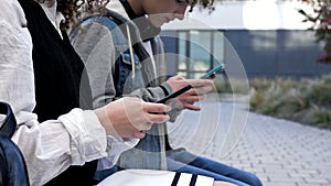 Two high school students, a boy and a girl, are sitting on a bench in the school yard. Each teenager has their own