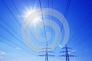 Two high-voltage pylons with high-voltage lines backlit by blue sky