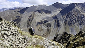 Two hikers are walking through difficult mountainous terrain on a cliff slope, with peaks of rocky mountains visible in