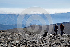 Two hikers walking on slate ground descending from the top of Mulhacen, then a plain crossed by a path, then a mountain range and photo