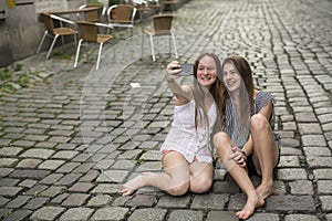 Two hilarious teenage girls are doing selfie on the phone sitting on the pavement.