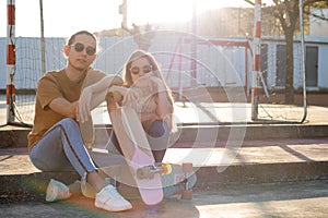 Two hipster friends sitting on steps leaning on cute skate board in a spring afeternoon with sunlight behind them