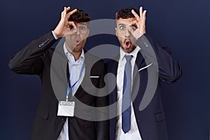 Two hispanic business men wearing business clothes doing ok gesture shocked with surprised face, eye looking through fingers