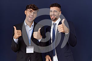 Two hispanic business men wearing business clothes success sign doing positive gesture with hand, thumbs up smiling and happy