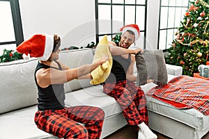 Two hispanic men couple fighting with pillow sitting by christmas tree at home