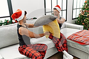 Two hispanic men couple fighting with pillow sitting by christmas tree at home