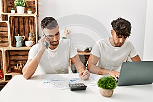 Two hispanic men couple talking on the smartphone and using laptop working at home