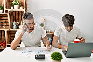 Two hispanic men couple talking on the smartphone and using laptop working at home