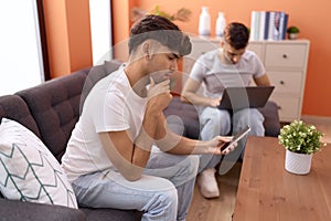 Two hispanic men couple using laptop and smartphone sitting on sofa at home