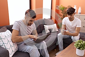 Two hispanic men couple using laptop and smartphone sitting on sofa at home