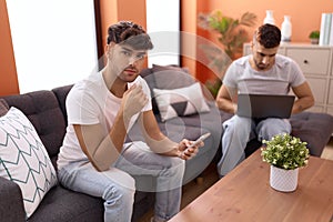 Two hispanic men couple using laptop and smartphone sitting on sofa at home