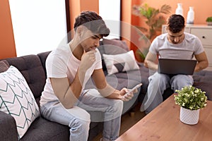 Two hispanic men couple using laptop and smartphone sitting on sofa at home