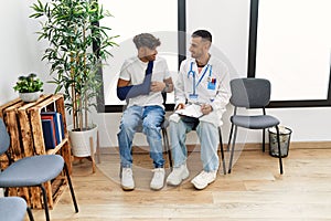 Two hispanic men doctor and patient writing on clipboard sitting on chair at hospital waiting room