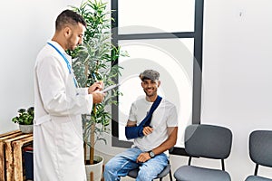 Two hispanic men doctor and patient writing on clipboard sitting on chair at hospital waiting room