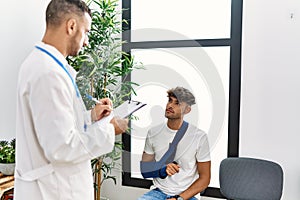 Two hispanic men doctor and patient writing on clipboard sitting on chair at hospital waiting room