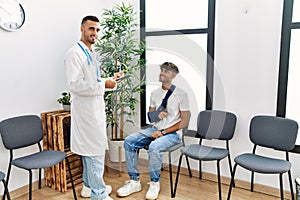 Two hispanic men doctor and patient writing on clipboard sitting on chair at hospital waiting room