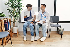 Two hispanic men doctor and patient writing on clipboard sitting on chair at hospital waiting room