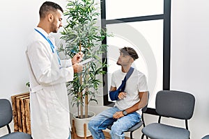 Two hispanic men doctor and patient writing on clipboard sitting on chair at hospital waiting room