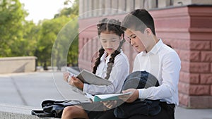 Two hispnic children students pupils sitting outdoors with books reading after class. Girl reads book sad boy showing