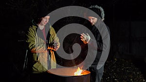 Two homeless young men bask in the fire. Men stand near a barrel of fire at night.