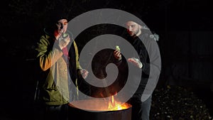 Two homeless young men are frying apples on a fire. Men stand at night near a barrel of fire and bask.