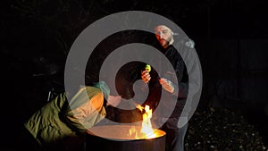 Two homeless young men stand by the fire and eat apples. Men stand at night near a barrel of fire and bask.