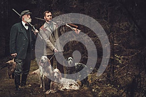 Two hunters with dogs and shotguns in a traditional shooting clothing, posing on a dark forest background.