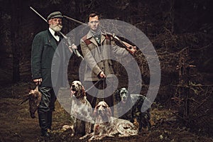 Two hunters with dogs and shotguns in a traditional shooting clothing, posing on a dark forest background.