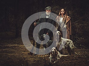 Two hunters with dogs and shotguns in a traditional shooting clothing, posing on a dark forest background.