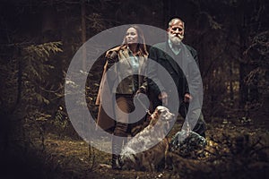 Two hunters with dogs and shotguns in a traditional shooting clothing, posing on a dark forest background.