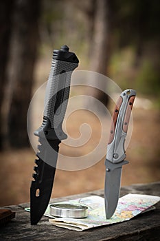 Two hunting knives are stuck in lid wooden table against background forest. Concept of adventure.